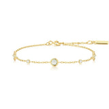 A delicate gold chain bracelet featuring a central round bezel-set moonstone and small clear accent stones, displayed on a clean white background. The brand name 'ANNA VAILI' is visible on a small gold tag at the clasp.